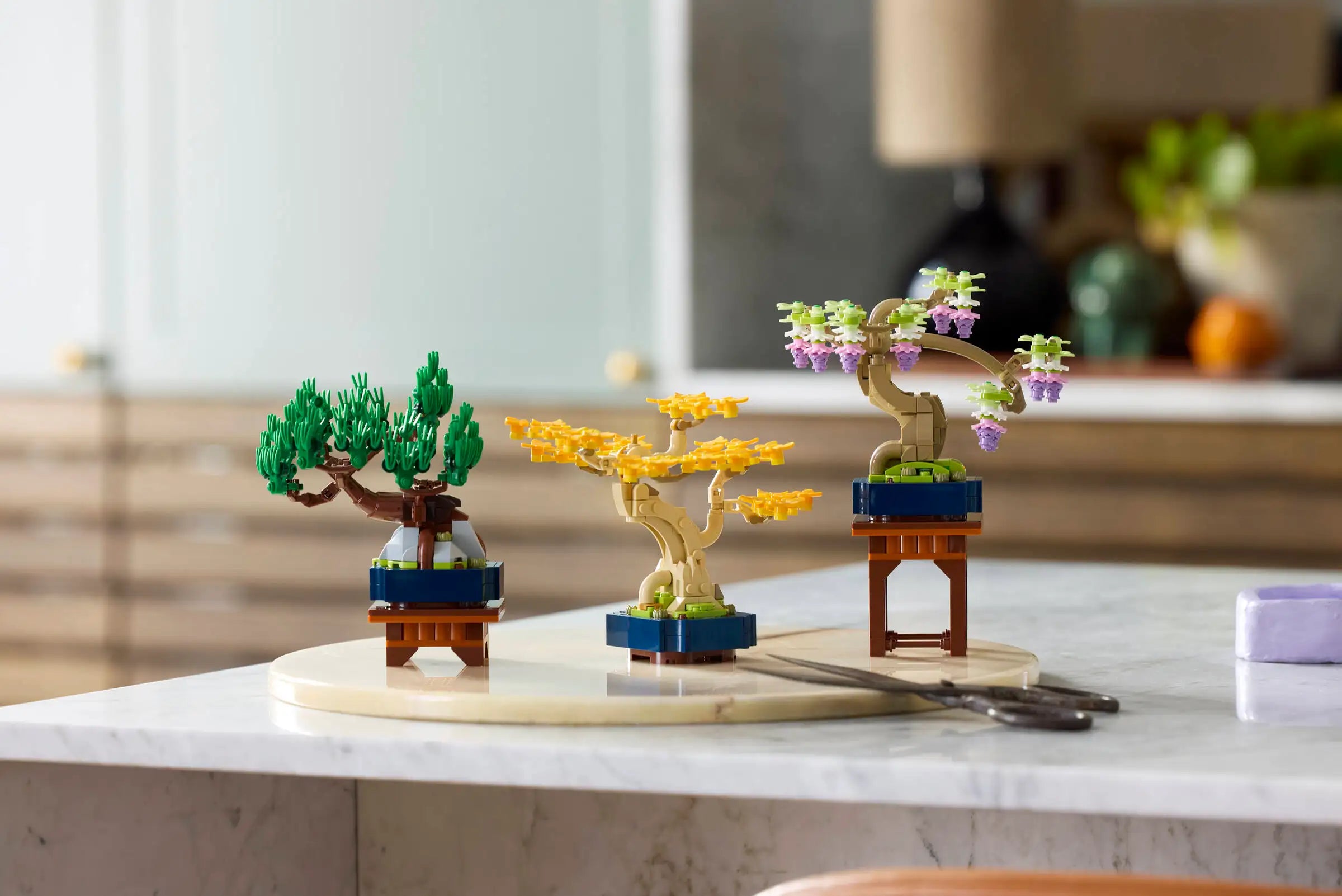 Three LEGO models of bonsai trees on a marble surface with a blurred indoor background.