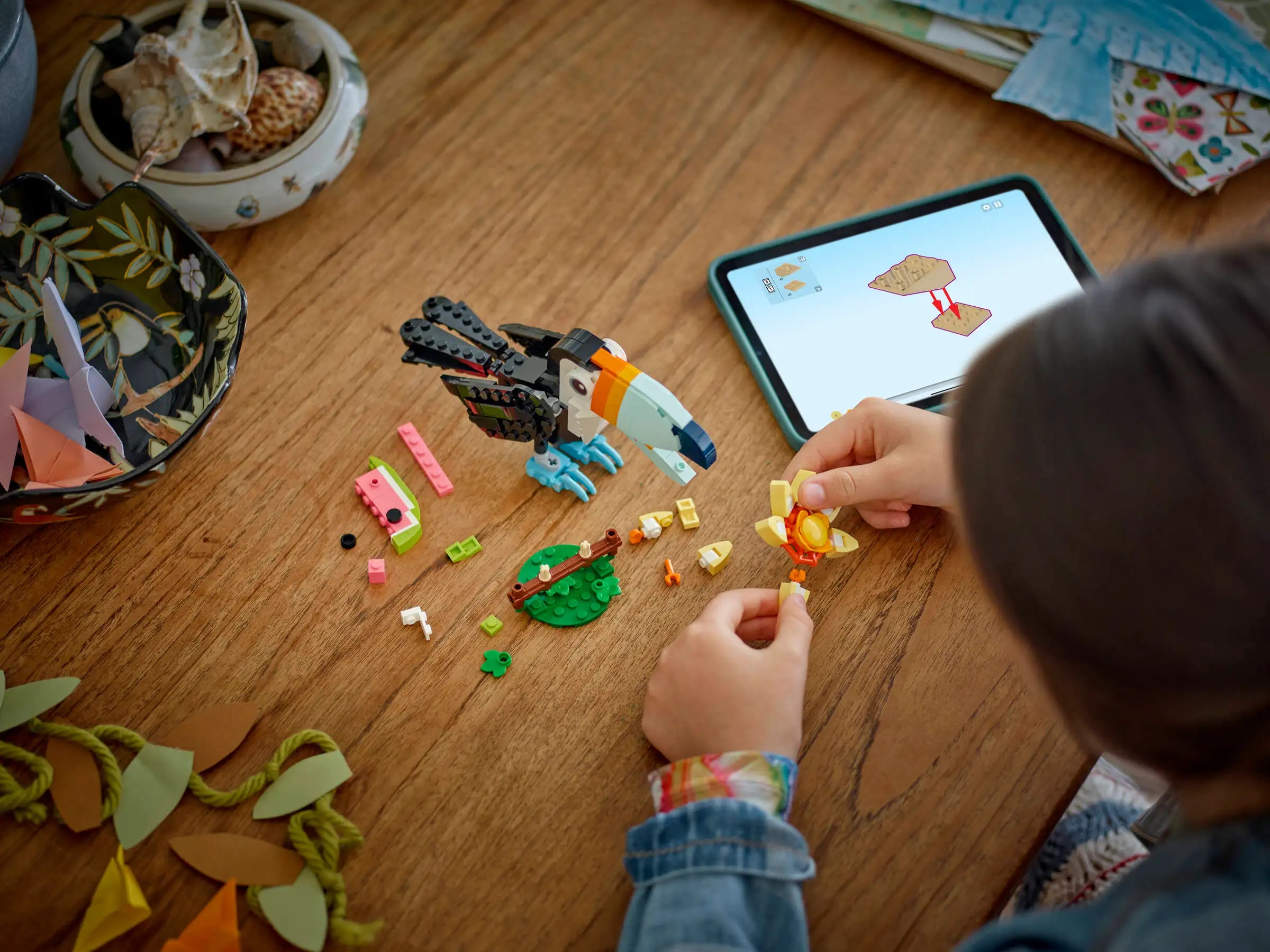 Child playing with LEGO 31173 Tropical Toucan set on a wooden table next to a tablet displaying a LEGO instruction app.