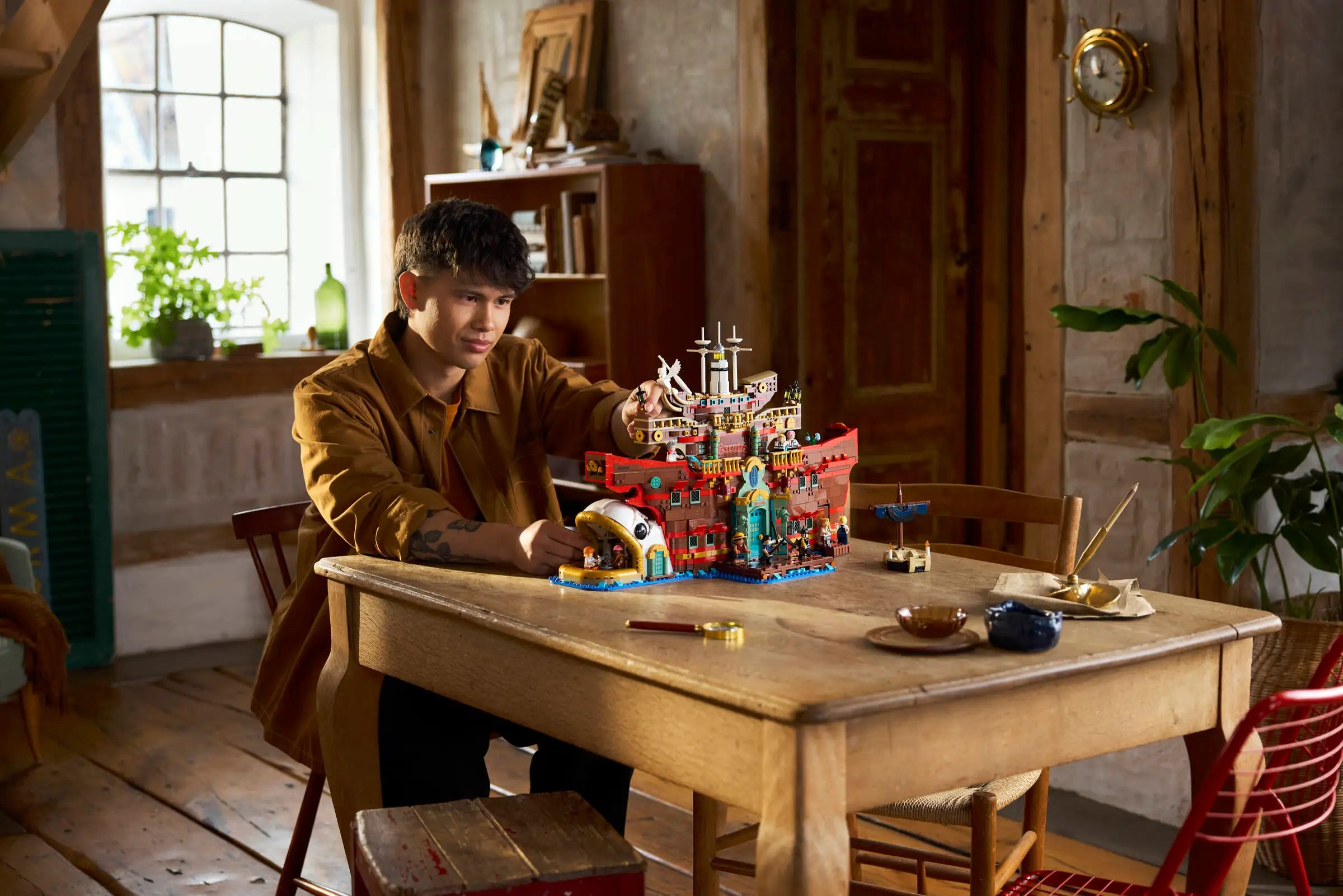 Person playing with LEGO 75640 The Baratie Floating Restaurant at a wooden table in a room with plants and furniture.