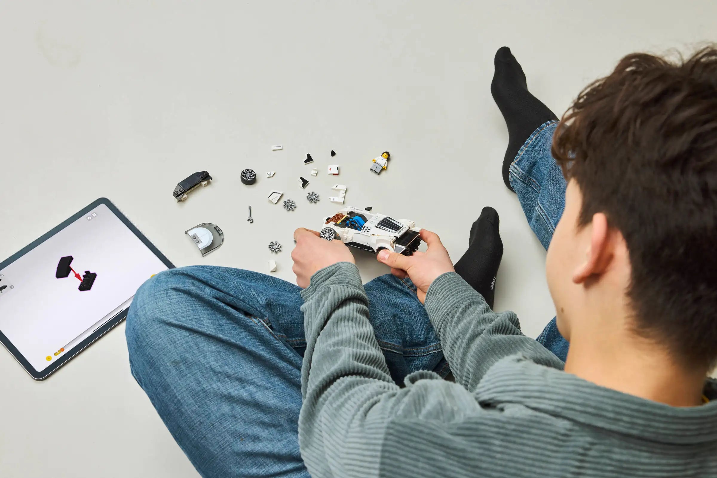 Person assembling a LEGO 77240 Bugatti Centodieci model car with parts on the floor, using a tablet for reference.