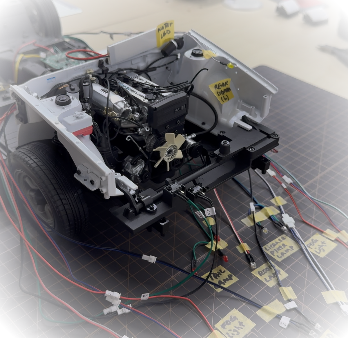 A white and black custom-built electric vehicle chassis with a visible internal engine and numerous attached wires, photographed on a gray cutting mat with handwritten yellow labels. - AE86 Current Build Status Transparent