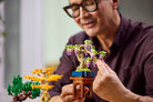Man holding a LEGO 10373 Mini Bonsai Trees of a tree with flowers, surrounded by additional models on a blurred background.