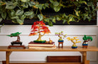 Collection of colorful bonsai tree models on a wooden table with a white wall and green plants in the background.